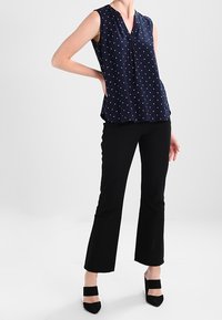 Kvinna iklädd en marinblå ärmlös blus med vita prickar, svarta figurnära byxor och svarta klackar med breda remmar, stående med en hand på höften.