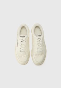 Hvite lær sneakers med perforert tå, flate snøringer, og minimal merkevare på siden og tungen. Gummisåle og polstret krage.