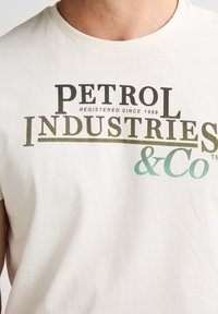 Camiseta blanca de algodón con un texto llamativo y multicolor: "PETROL INDUSTRIES & Co" con "registrado desde 1989" en una impresión más pequeña.