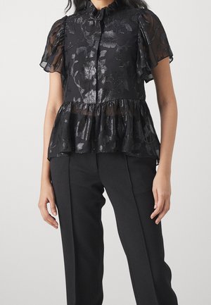 Vrouw draagt een zwarte doorschijnende bloemenblouse met geplooide korte mouwen en een peplumzoom, gecombineerd met op maat gemaakte zwarte broek.