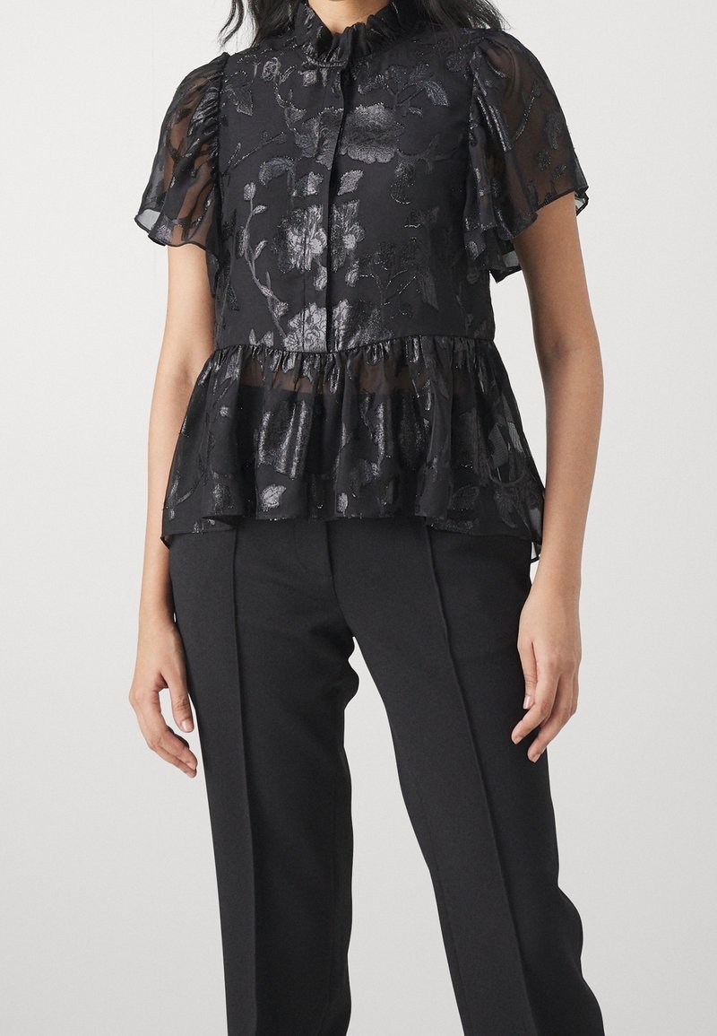 Femme portant un chemisier noir transparent à motifs floraux avec des manches courtes à volants et un basque, associé à un pantalon noir ajusté.
