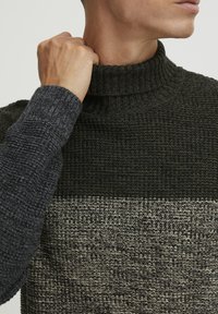 Teksturert strikket genser med høyt krage. Fargede blokker i mørkegrønn, grå og beige. Ribbede detaljer på kragen og mansjettene.