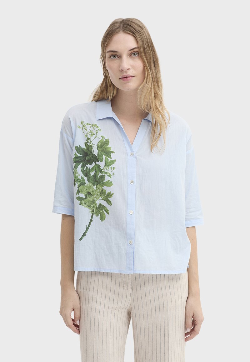 Femme portant une chemise bleu clair à boutons avec un imprimé botanique vert sur le côté gauche et un pantalon beige à fines rayures, debout devant un fond uni.