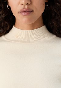 Maglione a collo alto a coste in crema, caratterizzato da un design a maglia testurizzata con una finitura liscia e accenti di stile minimalista e classico.