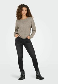 Jonge vrouw met krullend haar, gekleed in een grijs trui, zwarte skinny jeans en zwarte leren enkellaarsjes, staande met haar handen in haar zakken.