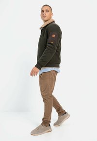 Dark green sweater jacket with a beige collar, brown sleeve patch, paired with light blue shirt and beige pants, finished with gray sneakers.