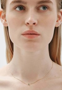 Collar de cadena de oro con un diseño minimalista, que presenta un pequeño acento de piedra verde, llevado alrededor de un cuello desnudo. Textura suave, forma delicada.