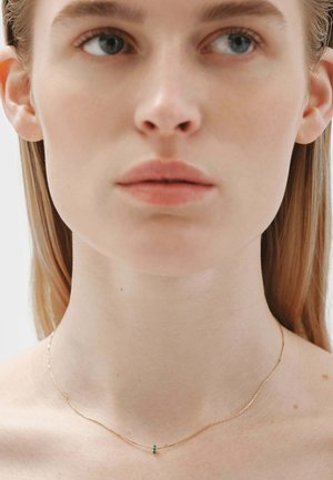 Collar de cadena de oro con un diseño minimalista, que presenta un pequeño acento de piedra verde, llevado alrededor de un cuello desnudo. Textura suave, forma delicada.