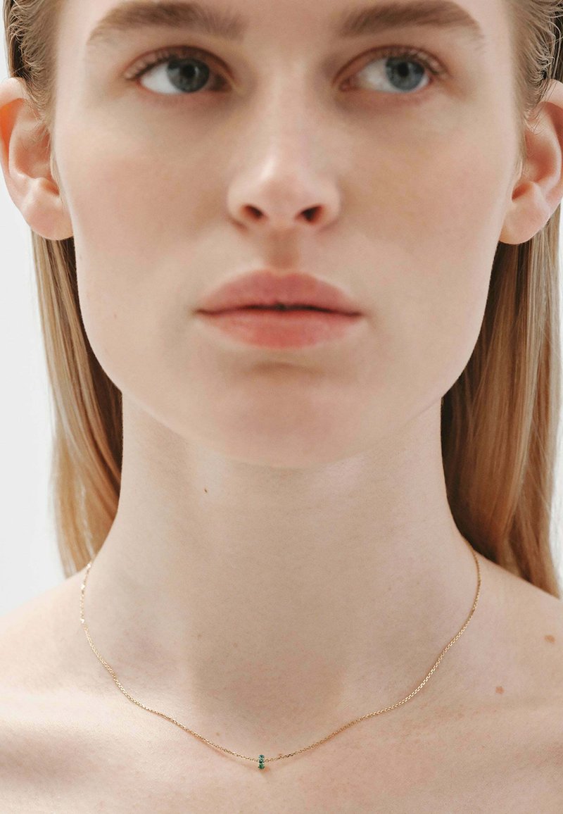 Collar de cadena de oro con un diseño minimalista, que presenta un pequeño acento de piedra verde, llevado alrededor de un cuello desnudo. Textura suave, forma delicada.