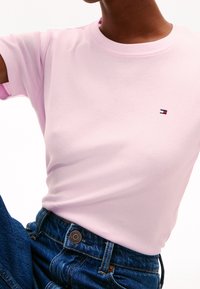 Růžové bavlněné tričko s kulatým výstřihem a krátkými rukávy, s malou vyšitou logem na hrudi. Kombinováno s modrými džínami.