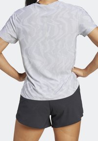 Camisa atlética de color gris claro con un patrón de zigzag texturizado, mangas cortas y una pequeña abertura en la parte posterior, combinada con pantalones cortos negros.