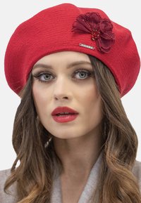 Beret roșu confecționat dintr-un material texturat, cu un accent floral închis, mărgele și un detaliu metalic cu logo. Formă netedă, rotundă.