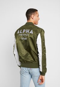 Olivgrüne Bomberjacke mit weißen Seitenpanelen, gerippten Bündchen und einem großen, gestickten Logo auf dem Rücken. Glattes, leichtes Material.