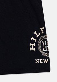 Zwart katoenen T-shirt met een crèmekleurige graphic op de linksonder, waarop de woorden "HILFIGER" en "NEW" in een circulair ontwerp staan.