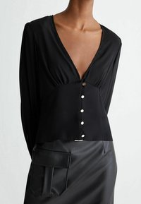 Donna che indossa una blusa nera con scollatura a V profonda e bottoni, insieme a una gonna di pelle nera con una grande tasca rettangolare sul lato.