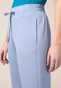 Jogging léger bleu en tissu doux avec une taille élastique à cordon. Présente un brodé "SOFT LIFE" et des poches latérales. Texture lisse.