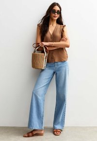 Haut sans manches brun à boutons avec des manches volantes, jean large bleu clair, sandales à lanières marron et un sac tissé avec une bandoulière.