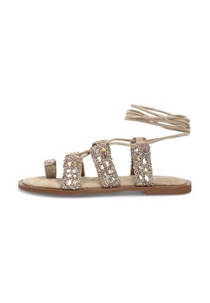 Sandalia plana beige con tiras adornadas con joyas, lazo para el dedo gordo y cintas para atar alrededor del tobillo sobre una suela marrón.