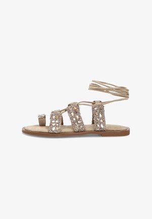 Sandalia plana beige con tiras adornadas con joyas, lazo para el dedo gordo y cintas para atar alrededor del tobillo sobre una suela marrón.