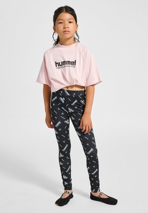 T-shirt coupe courte rose avec le logo "hummel!", associée à des leggings noirs avec des motifs graphiques blancs, et des chaussures plates noires avec un détail rayé.