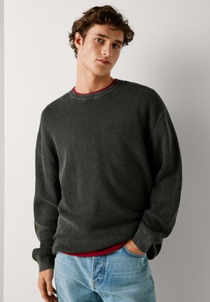 Donkergrijze gebreide trui met een ronde halslijn en geribbelde manchetten, gedragen over een rood shirt. Gecombineerd met lichtblauwe spijkerbroek.