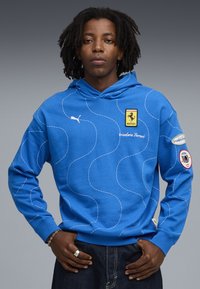 Felpa blu realizzata in tessuto morbido, con motivi a onda bianchi e logo Ferrari. Include un logo Puma sulla parte anteriore e toppe con il marchio sulle maniche.