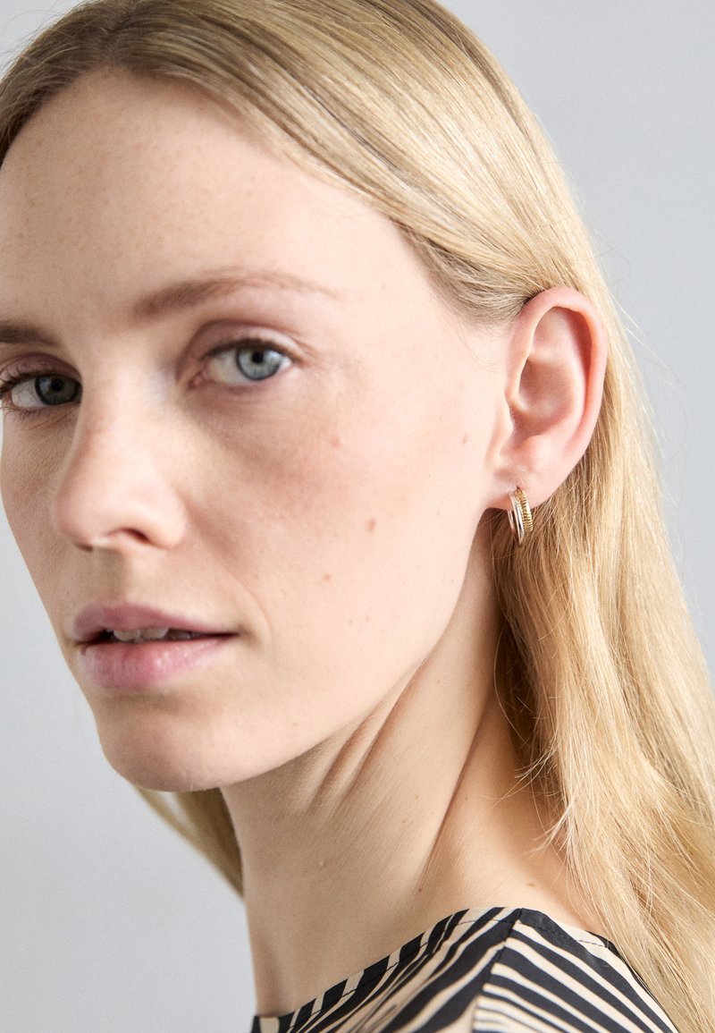 Zelta apli juvelierizstrādājumi ar teksturētu dizainu, ko valkā modele ar taisniem gaiši blondiem matiem. Fons ir maigs un neitrāls.