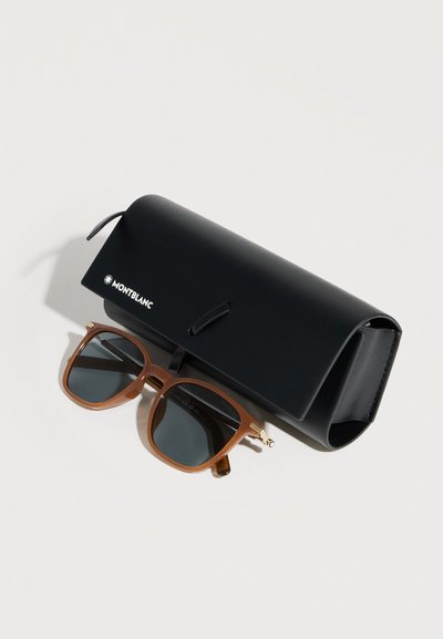 Γυαλιά ηλίου με καφέ κορνίζα και σκουρούς φακούς τοποθετημένα δίπλα σε μαύρη δερμάτινη θήκη γυαλιών ηλίου με λογότυπο Montblanc πάνω σε λευκή επιφάνεια.