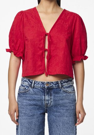 Rød kort bluse med pufærmer og knyttelukninger foran, båret med højtaljede blå jeans med syrevask.