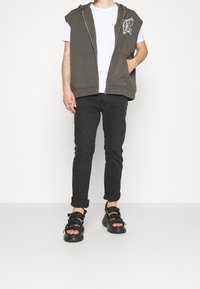 Hombre con sudadera sin mangas negra, camiseta blanca, vaqueros negros arremangados, sandalias negras con suelas gruesas y anillos en la mano izquierda.