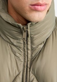 Olive green padded jacket with high collar, zip closure, and quilted pattern. Fabric has a smooth texture and subtle sheen.