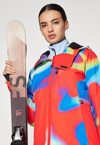 Suusajakk, millel on elavad punase, sinise ja rohelise mustrid, varustuses kapuuts ja lukuga tasku. Mudel hoiab suuski, millel on märge "Freestyle Park 50°."