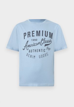 T-shirt en coton bleu clair avec un motif imprimé en bleu foncé, portant les inscriptions « Premium American Classic » et « Denim Goods ». Design à manches courtes.