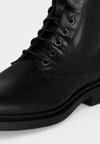 Gros plan d'une botte en cuir noir à lacets avec des œillets métalliques et des détails cousus sur le côté et à l'avant.