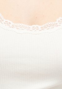 Κοντινό πλάνο ενός λευκού ριμπ αμάνικου τοπ με λεπτή λευκή δαντέλα κατά μήκος του ντεκολτέ.