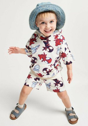 Smiling toddler wearing blue hat, Spiderman print hoodie and shorts, standing with legs apart and arms slightly outstretched on white background.