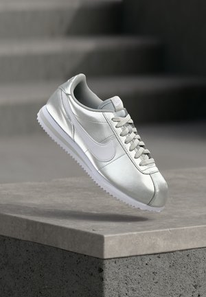 Ezüst Nike sportcipő fehér pipa és fűzőkkel, egy beton perem felett lebegve, háttérben elmosódott lépcsőkkel.