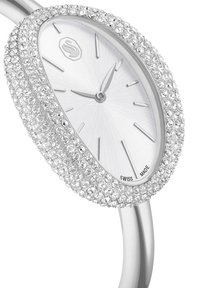 Zilveren horloge met een ovale kast bezet met kristallen, een witte, gestructureerde wijzerplaat en zilverkleurige wijzers; label "Swiss Made" nabij de onderkant.