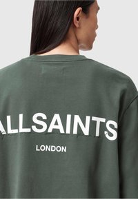 Zielona bluza z luźnym krojem. Posiada dużą białą grafikę "ALLSAINTS LONDON" oraz subtelne detale szycia na plecach. Materiał z mieszanki bawełny.