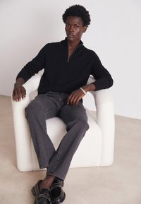Pull en tricot noir à fermeture éclair, jean gris et chaussures noires, assis sur une chaise blanche à texture avec bords arrondis, sur un fond neutre.
