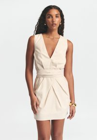 Béžové bezrukávové šaty s hlbokým výstrihom do V, zaväzovaním v páse a skladanou sukňou. Hladká textúra s voľným strihom a minimalistickým dizajnom.
