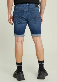 Shorts din denim de culoare albastră închisă, cu margini răsucite. Au două buzunare la spate cu cusături decorative. Se poartă cu șosete negre și adidași.