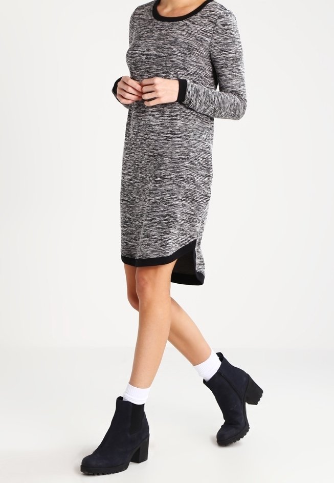 Femme portant une robe à manches longues chinée noir et blanc avec des bordures noires et des bottines noires avec des chaussettes blanches, posant en milieu de pas.