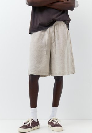 Person trägt beige Leinen-Shorts mit Kordelzug, weiße Socken und braune Low-Top-Sneaker, steht vor einem einfachen weißen Hintergrund.