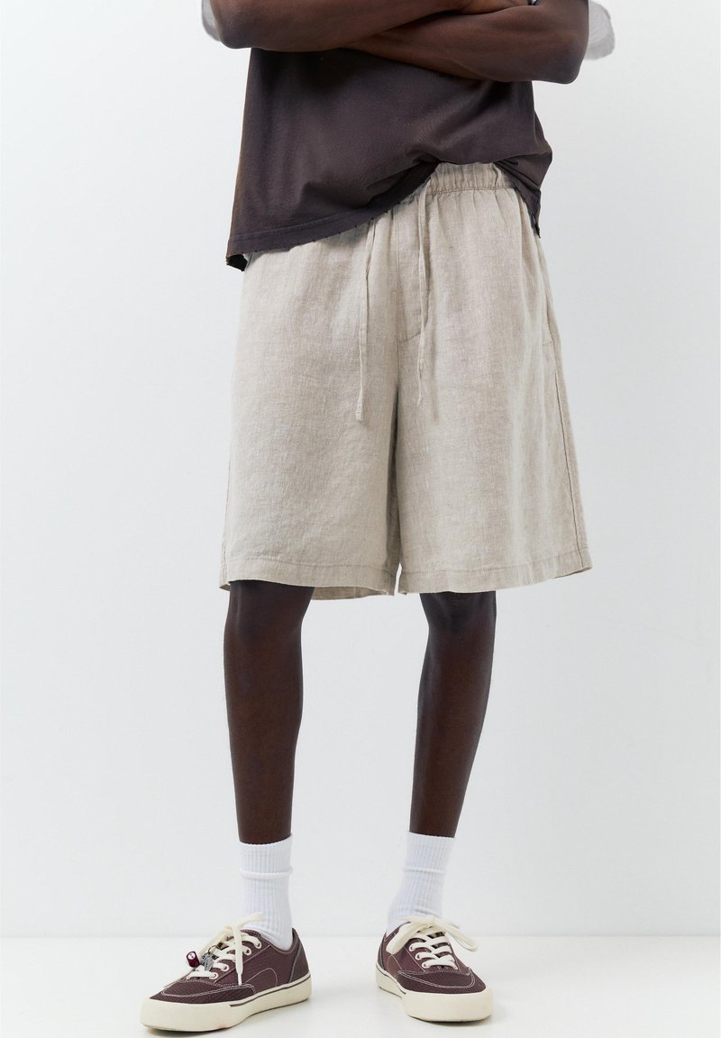 Personne portant un short beige en lin à cordon, des chaussettes blanches et des baskets basses marron, debout sur un fond blanc uni.