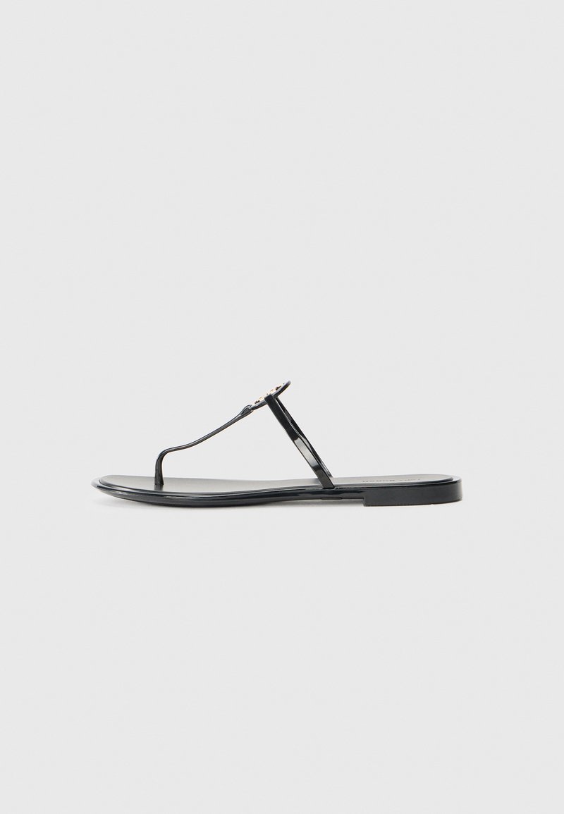 Svart flat sandal med tynne stropper og et lite dekorativt emblem på den sentrale stroppen, vist fra siden på en hvit bakgrunn.