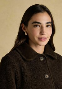 Hnedý, textúrovaný kabát s kordovou golierom, dvoma gombíkmi a jemným, nadýchaným vzhľadom. Svetlé pozadie zvýrazňuje zameranie na odev.