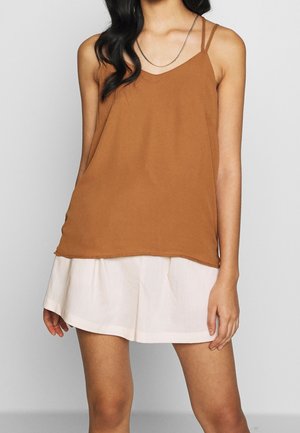 Femme portant un top marron sans manches avec de fines bretelles et une mini-jupe blanche, debout devant un fond gris.