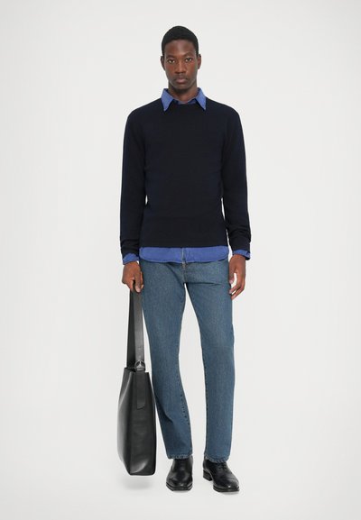 Marine gebreide trui, lichtblauw overhemd met kraag, blauwe spijkerbroek, zwarte leren tas met verstelbare schouderband en zwarte enkellaarsjes. Eenvoudige, casual look.