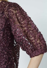 Burgunderrød kjole med transparent stof og tekstureret blomsterapplikation på ærmerne. Pufærmer med sammenrullede manchetter og indviklede detaljer.